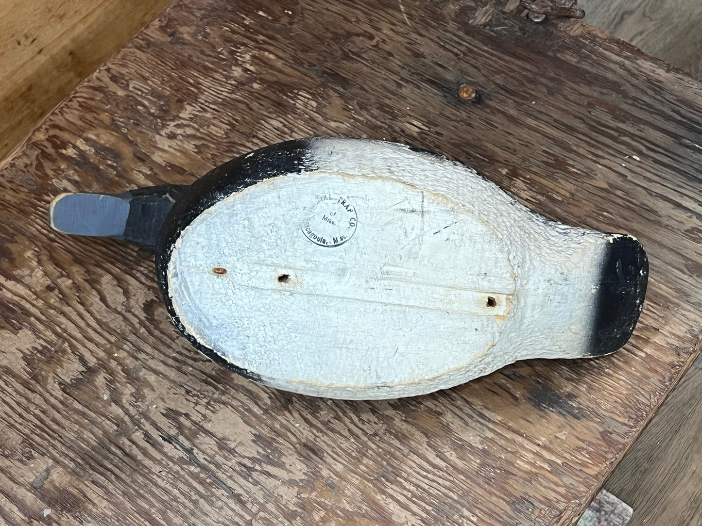A vintage wooden duck hunting blind or deco, shown from the underside on a rustic wooden table. The body is painted off-white with black tips on the tail and bill, and a gray plastic or metal blade is attached to the tail.