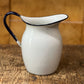 A white enamel pitcher with a dark blue rim and handle, set on a rustic wooden table against a wooden background.