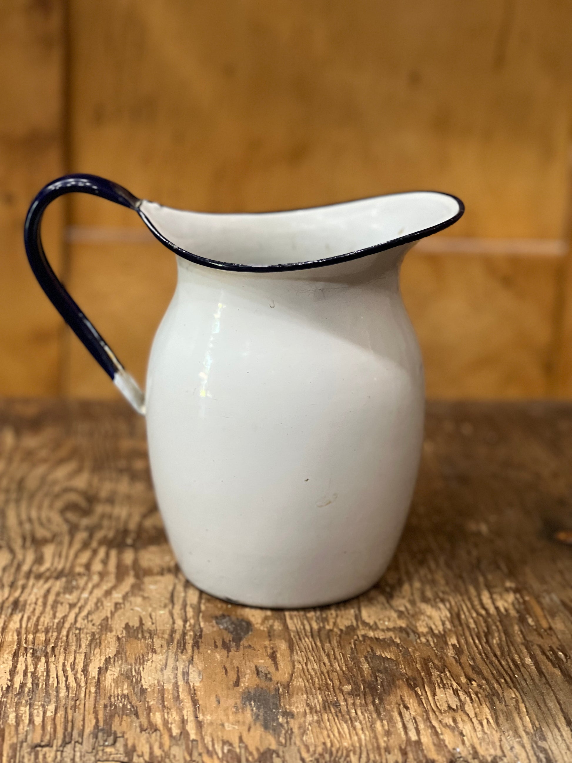 A white enamel pitcher with a dark blue rim and handle, set on a rustic wooden table against a wooden background.