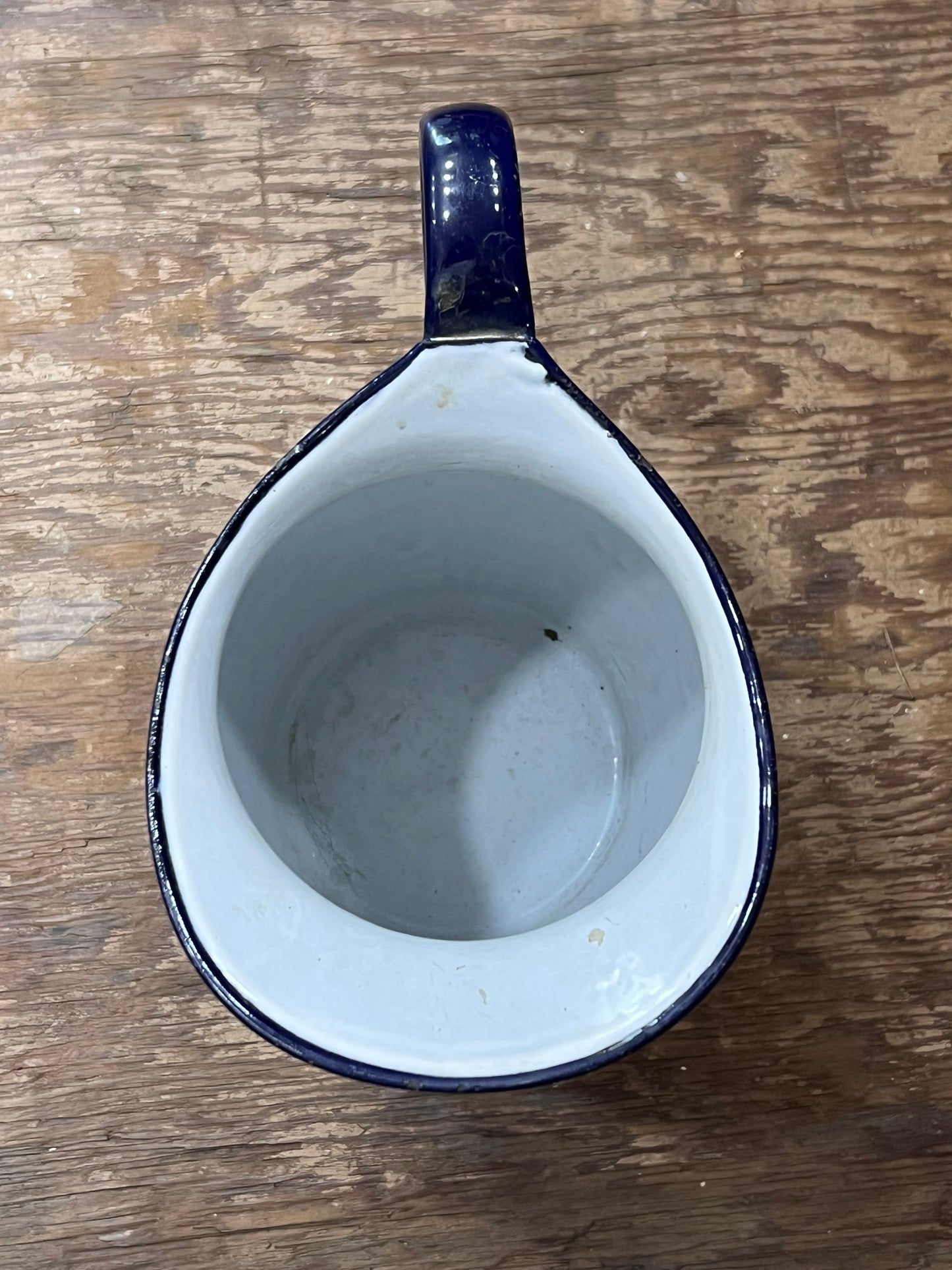 A vintage teardrop-shaped enamel pitcher with a dark blue exterior and white interior, shown from a top-down view on a rustic wooden surface.