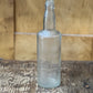 An empty, clear glass bottle with a narrow neck and thick body, standing on a rustic wooden surface with a wooden background.