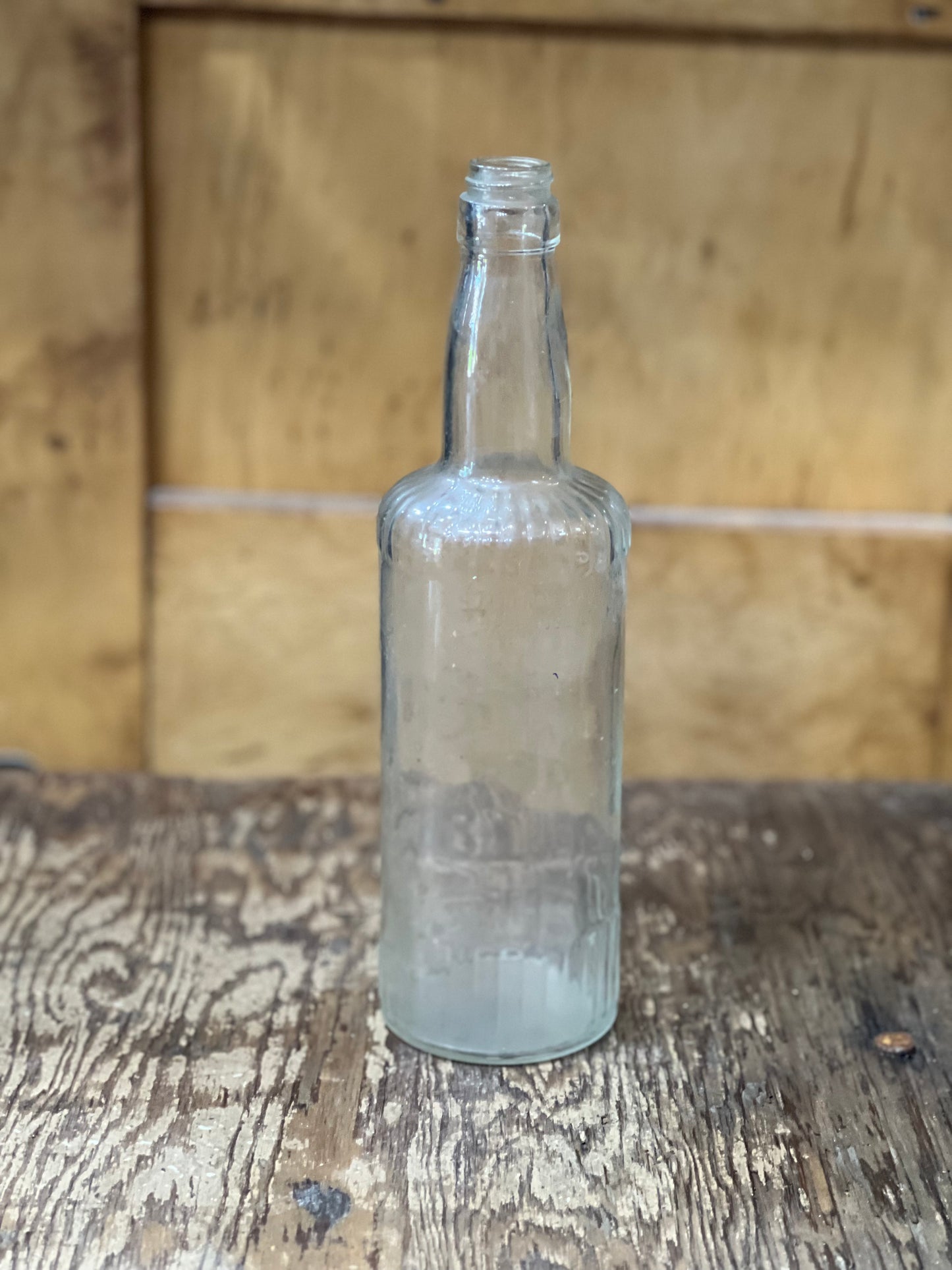 An empty, clear glass bottle with a narrow neck and thick body, standing on a rustic wooden surface with a wooden background.