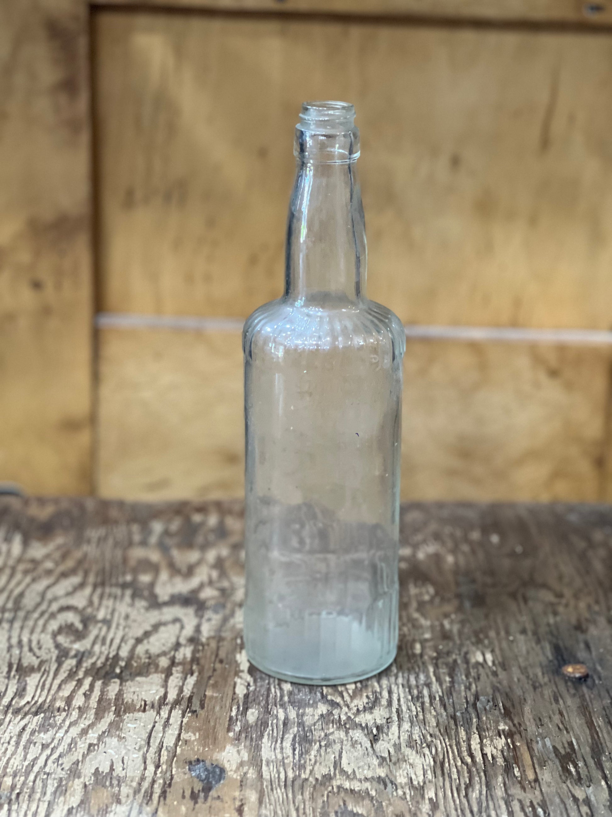 An empty, clear glass bottle with a narrow neck and thick body, standing on a rustic wooden surface with a wooden background.