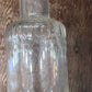 A clear vintage glass bottle with embossed text, shown against a rustic wooden plank background.