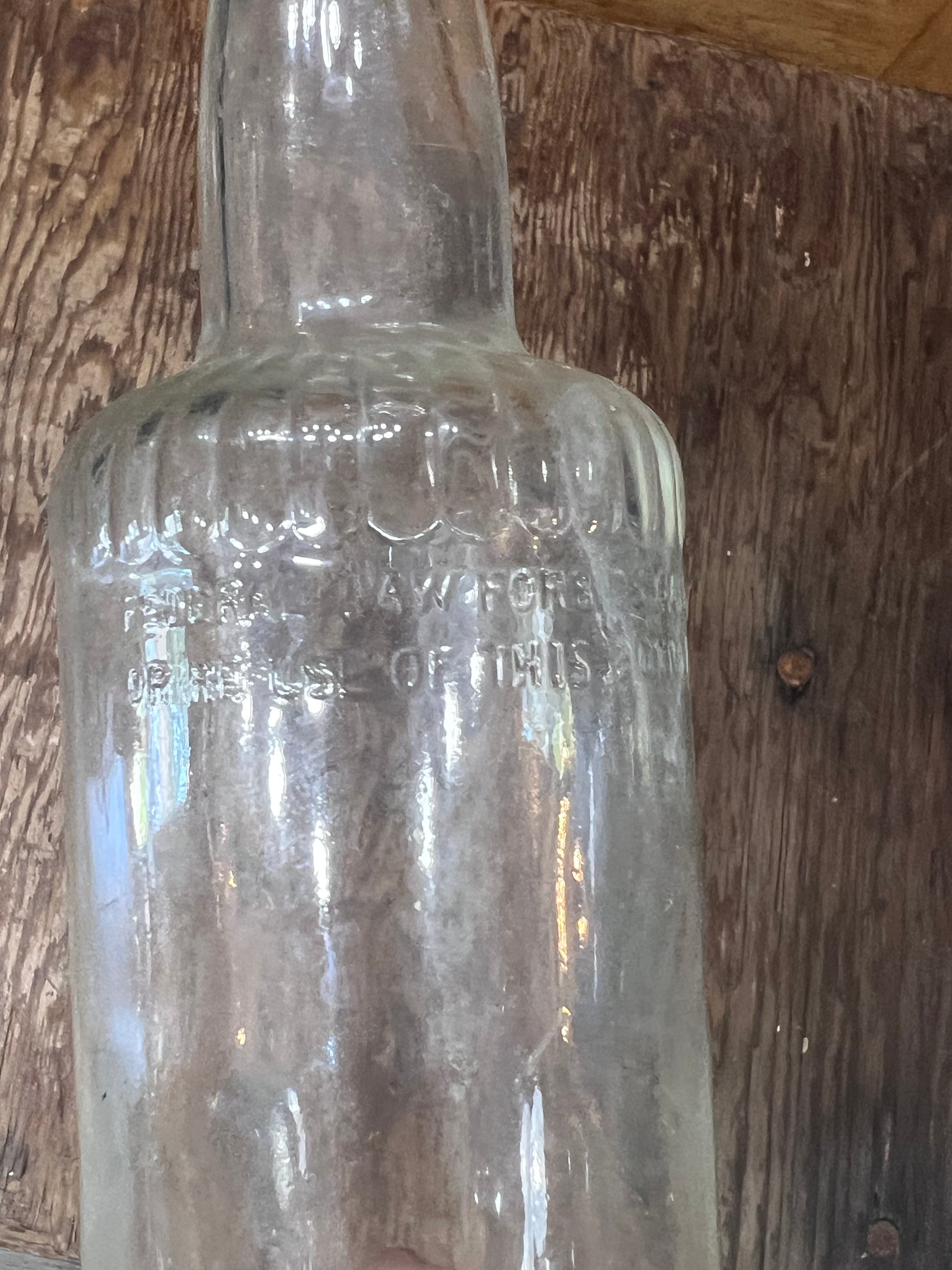 A clear vintage glass bottle with embossed text, shown against a rustic wooden plank background.