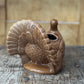 A brown glazed ceramic turkey-shaped planter with a textured feather pattern and a small opening at the top, shown on a rustic wooden surface with a blurred wood background.
