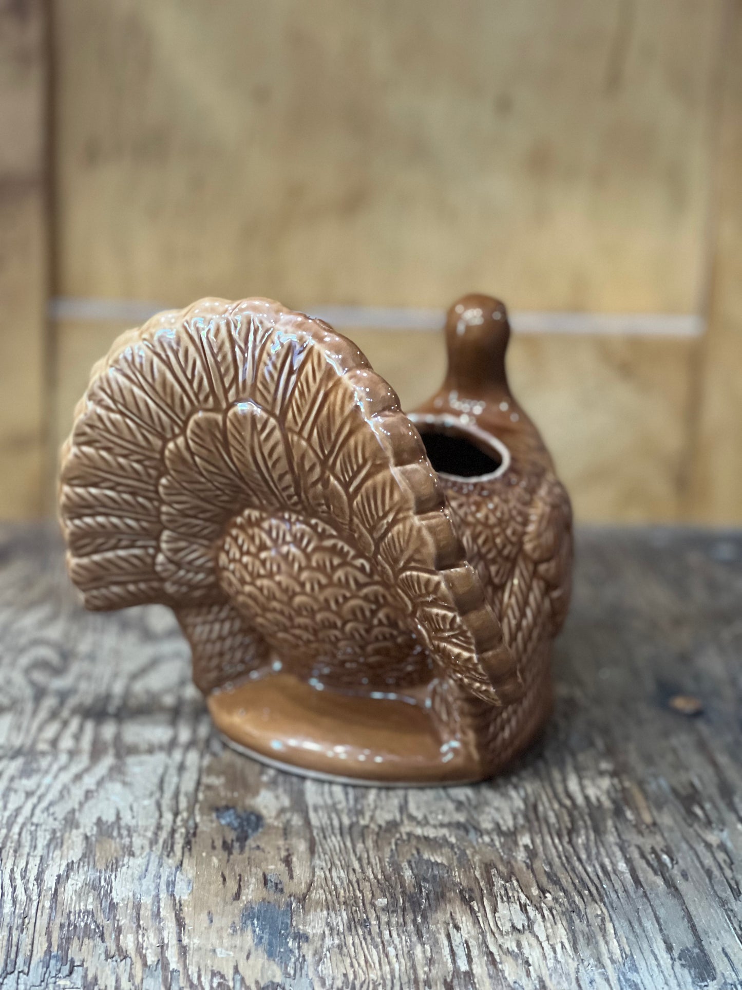 A brown glazed ceramic turkey-shaped planter with a textured feather pattern and a small opening at the top, shown on a rustic wooden surface with a blurred wood background.