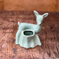 A small ceramic deer-shaped holder or cup in a pale mint green glaze, placed on a rustic wooden surface with a blurred brown background.