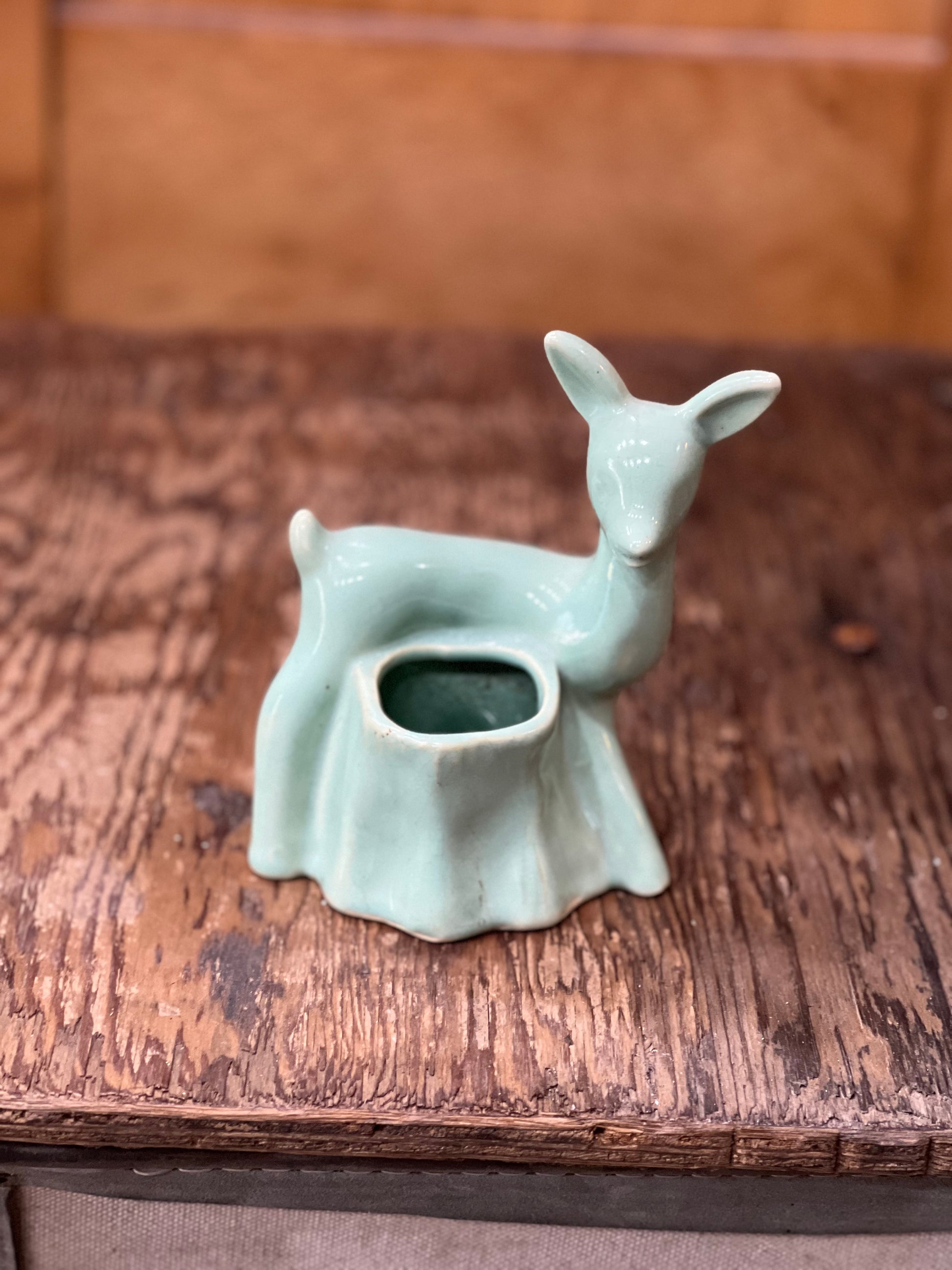 A small ceramic deer-shaped holder or cup in a pale mint green glaze, placed on a rustic wooden surface with a blurred brown background.