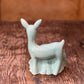 A small glossy ceramic deer figurine in a pale celadon green glaze, standing on a rustic dark wood surface with a blurred brown background.