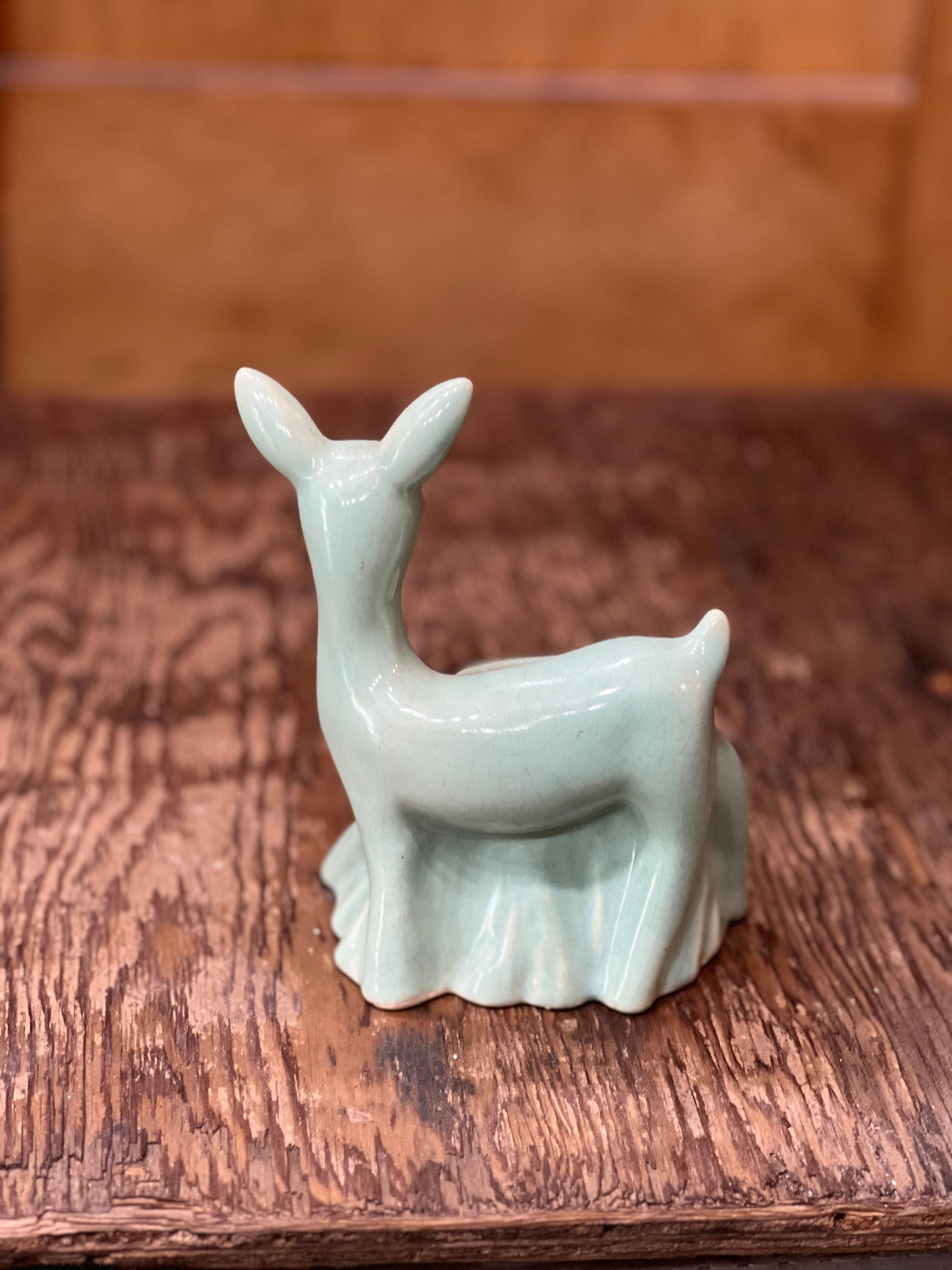 A small glossy ceramic deer figurine in a pale celadon green glaze, standing on a rustic dark wood surface with a blurred brown background.