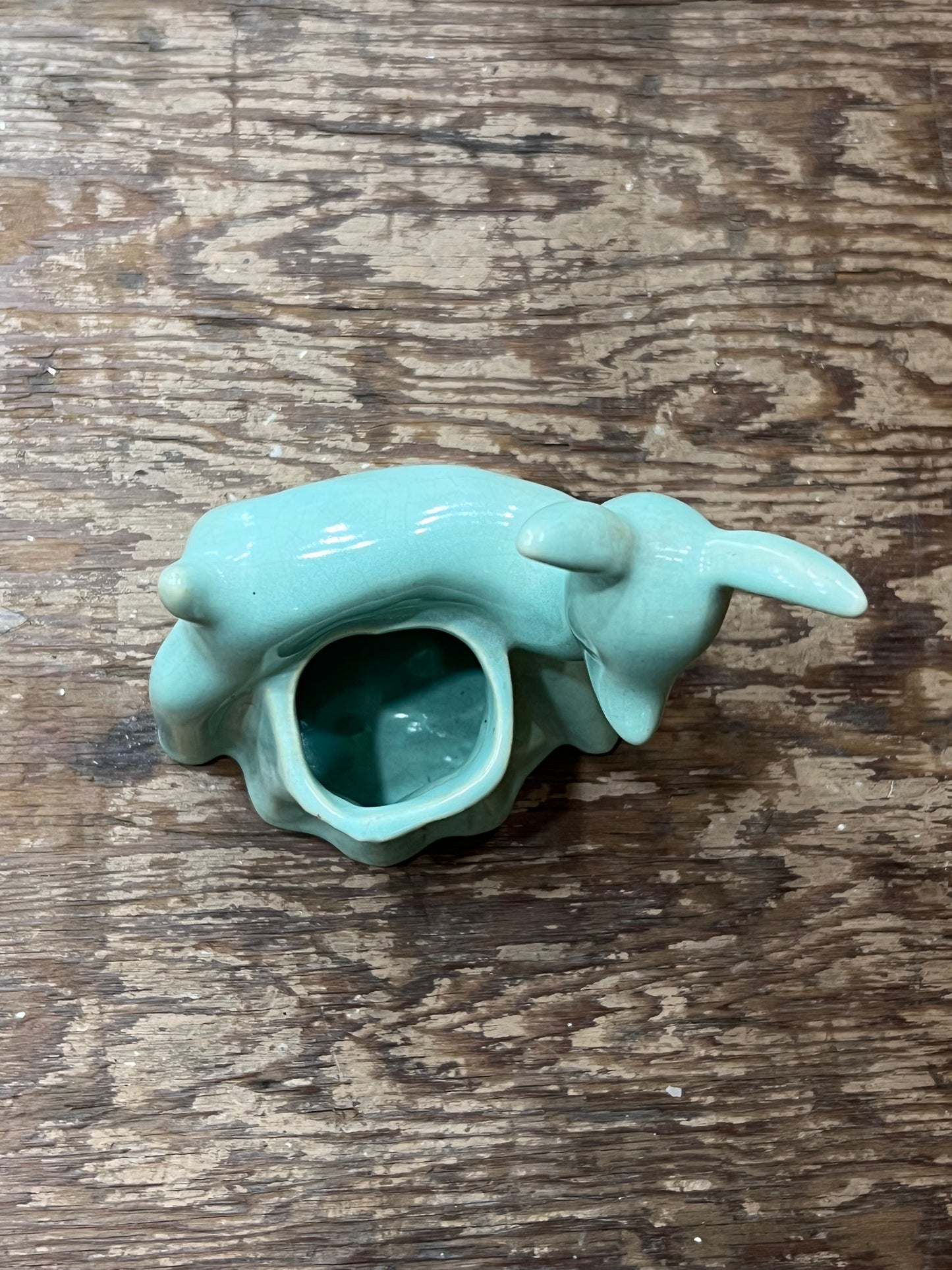 A small glossy ceramic animal-shaped vessel, shaped like a rabbit or hare with a hollow center, in a light aqua glaze, placed on a rustic wooden surface.