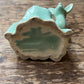 A small glossy ceramic deer figurine in a light aqua or mint green glaze, shown from the back on a rustic wooden tabletop. The base is unglazed, showing a natural beige clay texture.