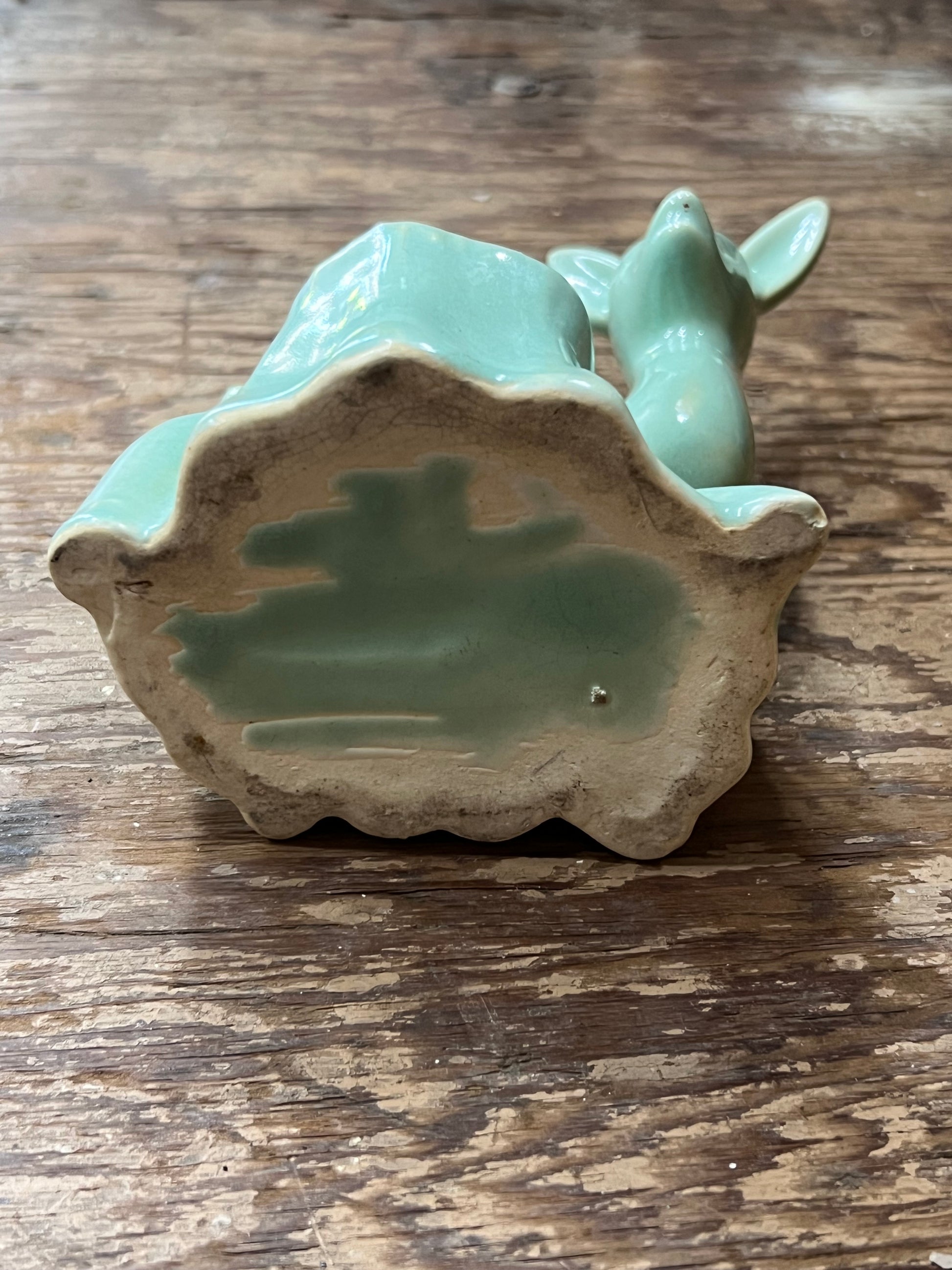 A small glossy ceramic deer figurine in a light aqua or mint green glaze, shown from the back on a rustic wooden tabletop. The base is unglazed, showing a natural beige clay texture.
