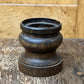 A dark brown carved wooden object with a flared top rim, resting on a rustic wooden surface against a light wood panel background.