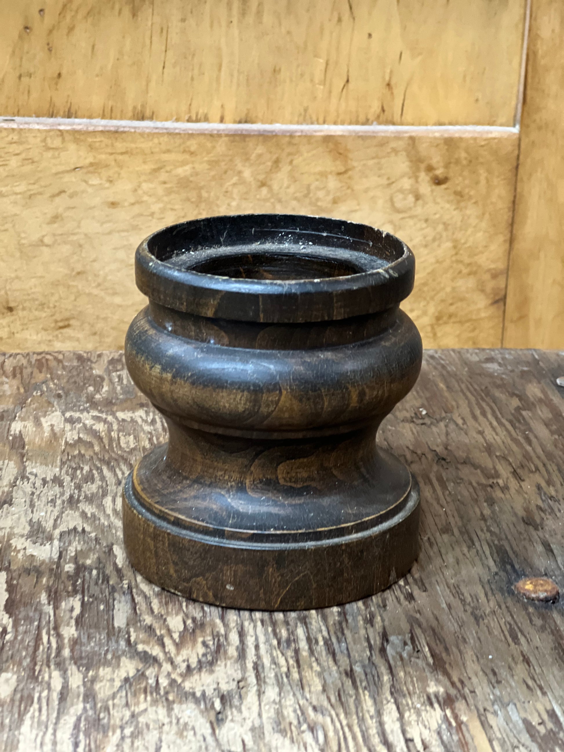 A dark brown carved wooden object with a flared top rim, resting on a rustic wooden surface against a light wood panel background.