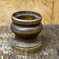 A vintage carved wooden candle holder or candle cup with a tiered base, shown on a rustic wooden table against a light wood paneled background.