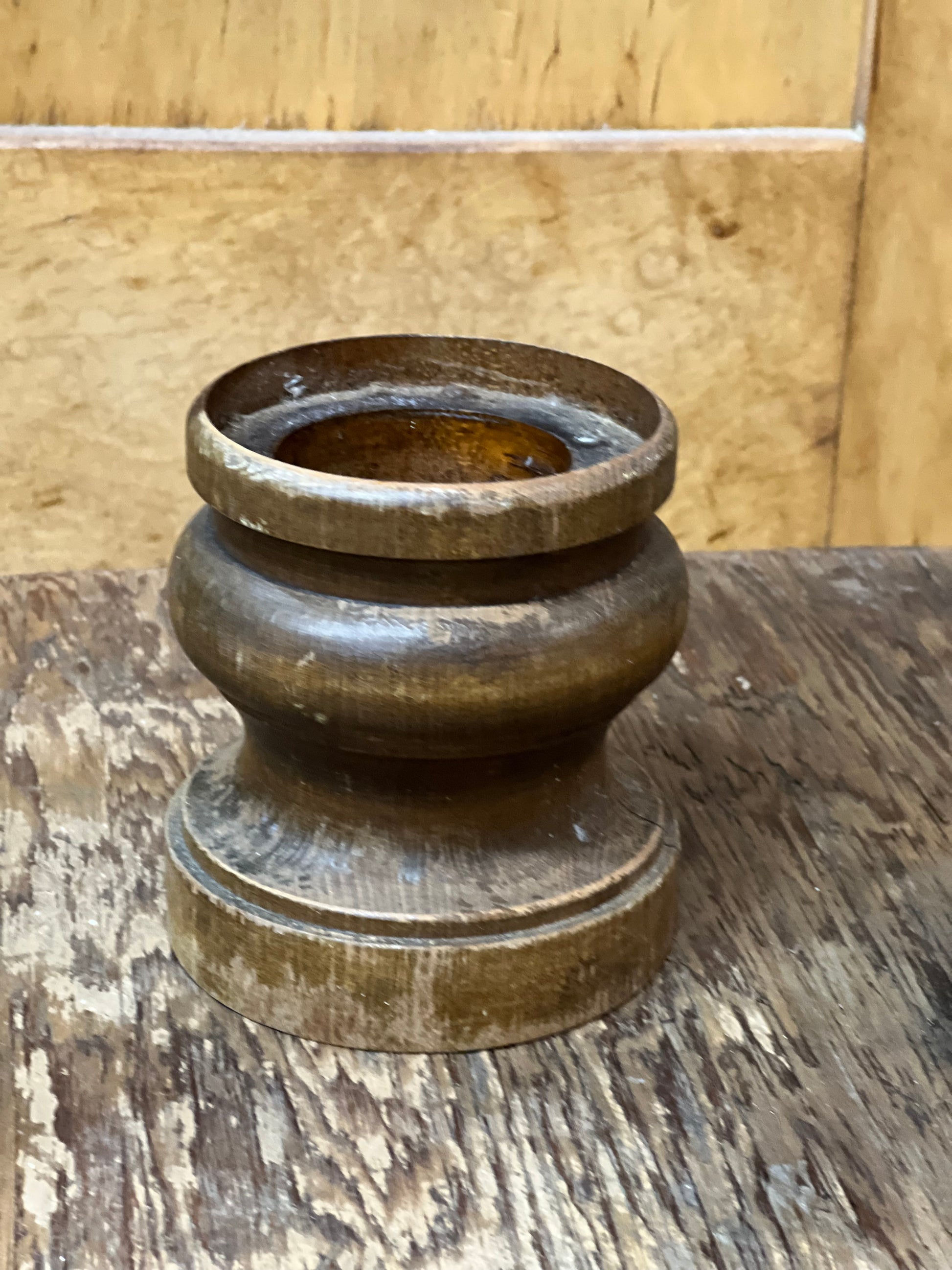 A vintage carved wooden candle holder or candle cup with a tiered base, shown on a rustic wooden table against a light wood paneled background.