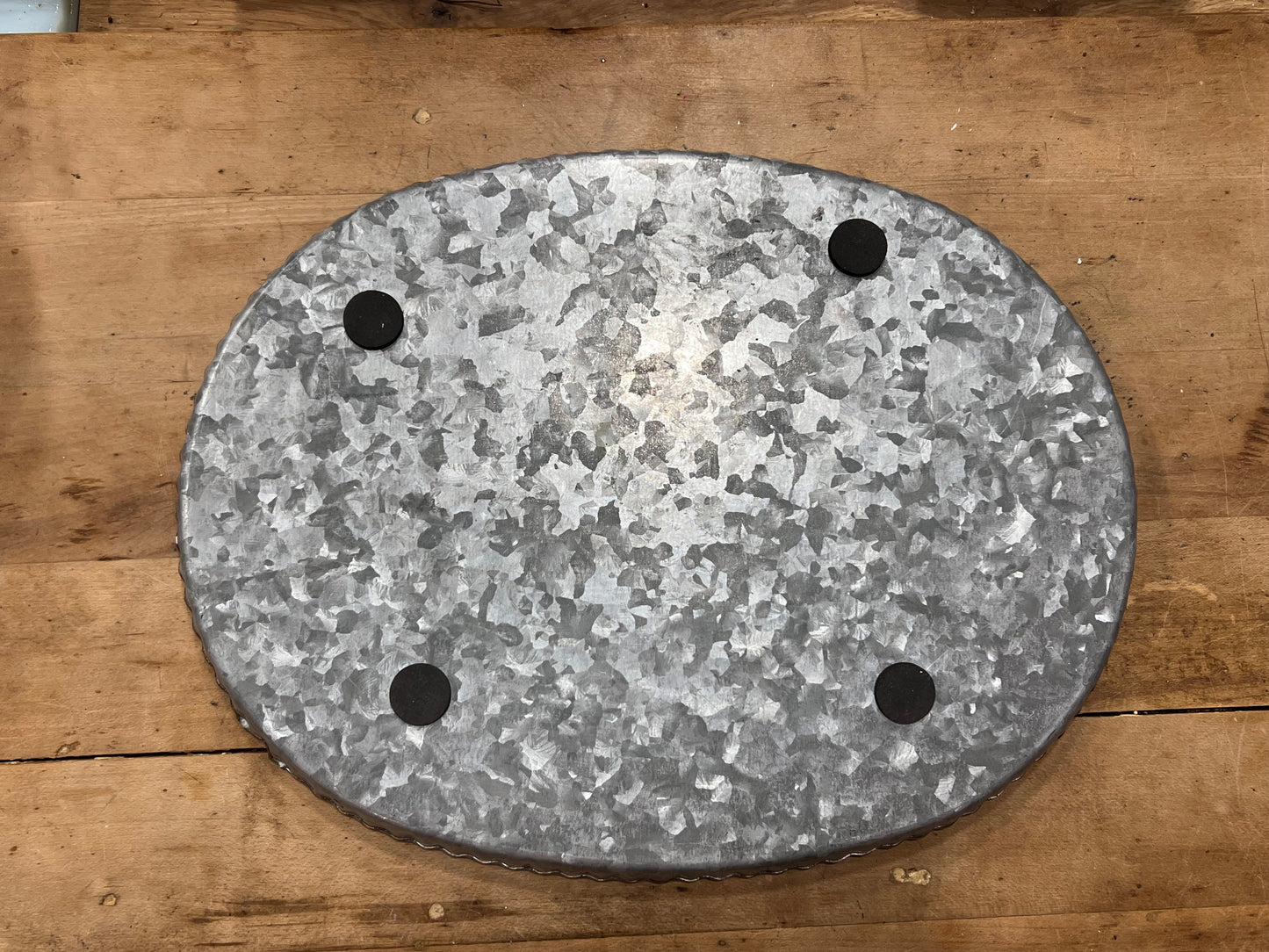 An oval galvanized metal stand or base with four circular feet, photographed from above on a rustic wooden surface.