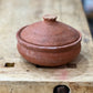 A small handmade terracotta clay lidded pot with a rounded body and matching lid, resting on a rustic wooden workbench.