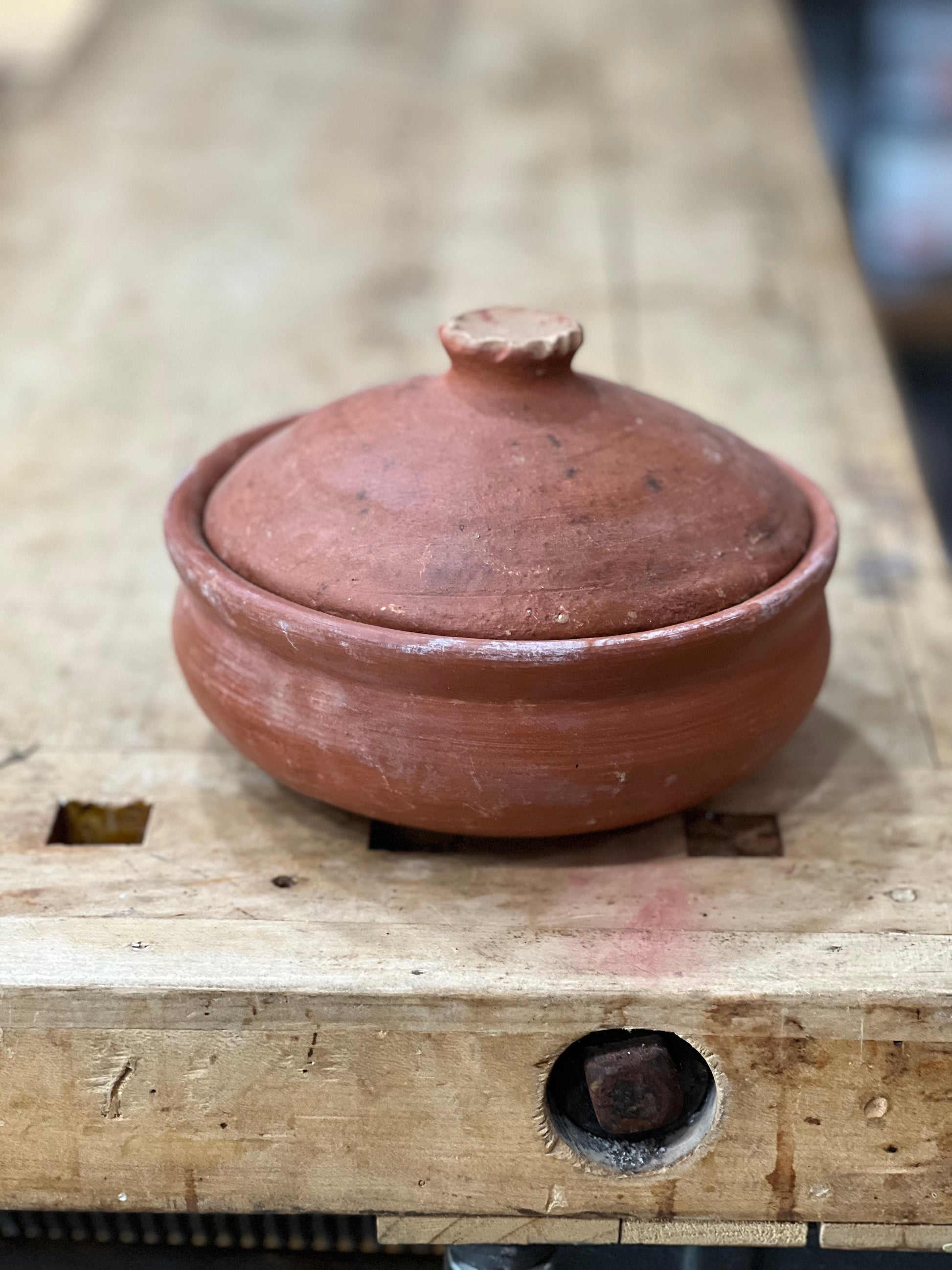 A small handmade terracotta clay lidded pot with a rounded body and matching lid, resting on a rustic wooden workbench.