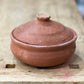 A small handmade terracotta clay lidded pot with a rounded body and matching lid, resting on a rustic wooden workbench.