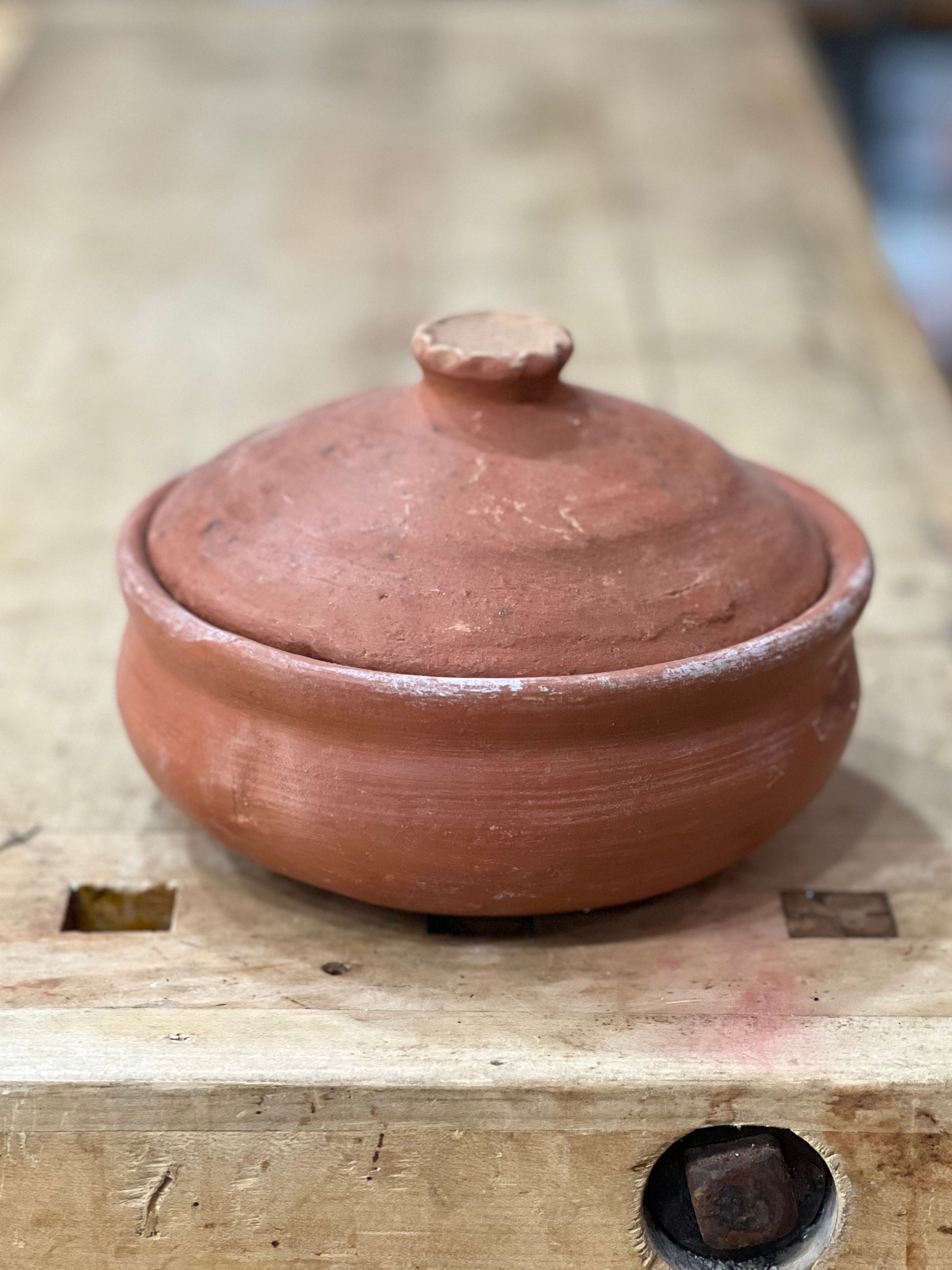 A small handmade terracotta clay lidded pot with a rounded body and matching lid, resting on a rustic wooden workbench.