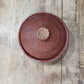 A round terracotta clay lidded pot or tureen shown from above on a rustic wooden plank surface.
