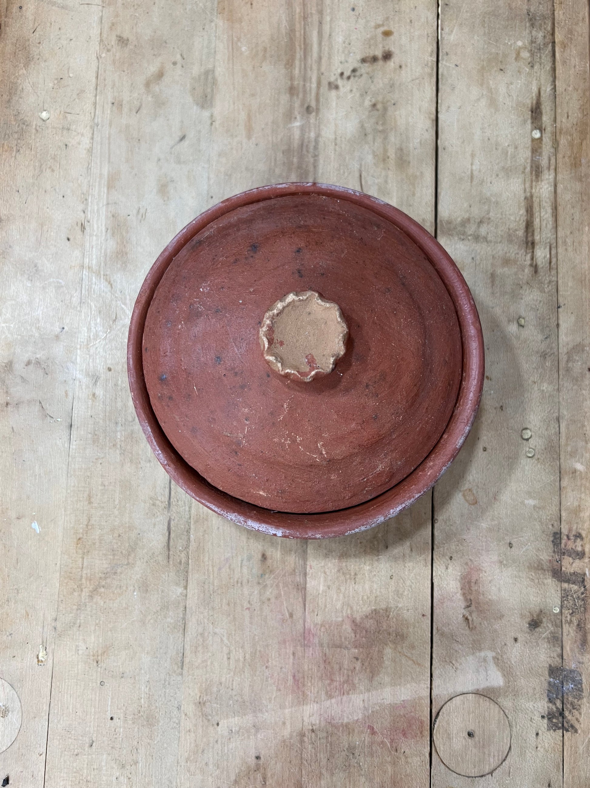 A round terracotta clay lidded pot or tureen shown from above on a rustic wooden plank surface.
