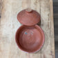A handmade terracotta clay lidded bowl with a matching lid resting on top, displayed on a rustic wooden plank surface.