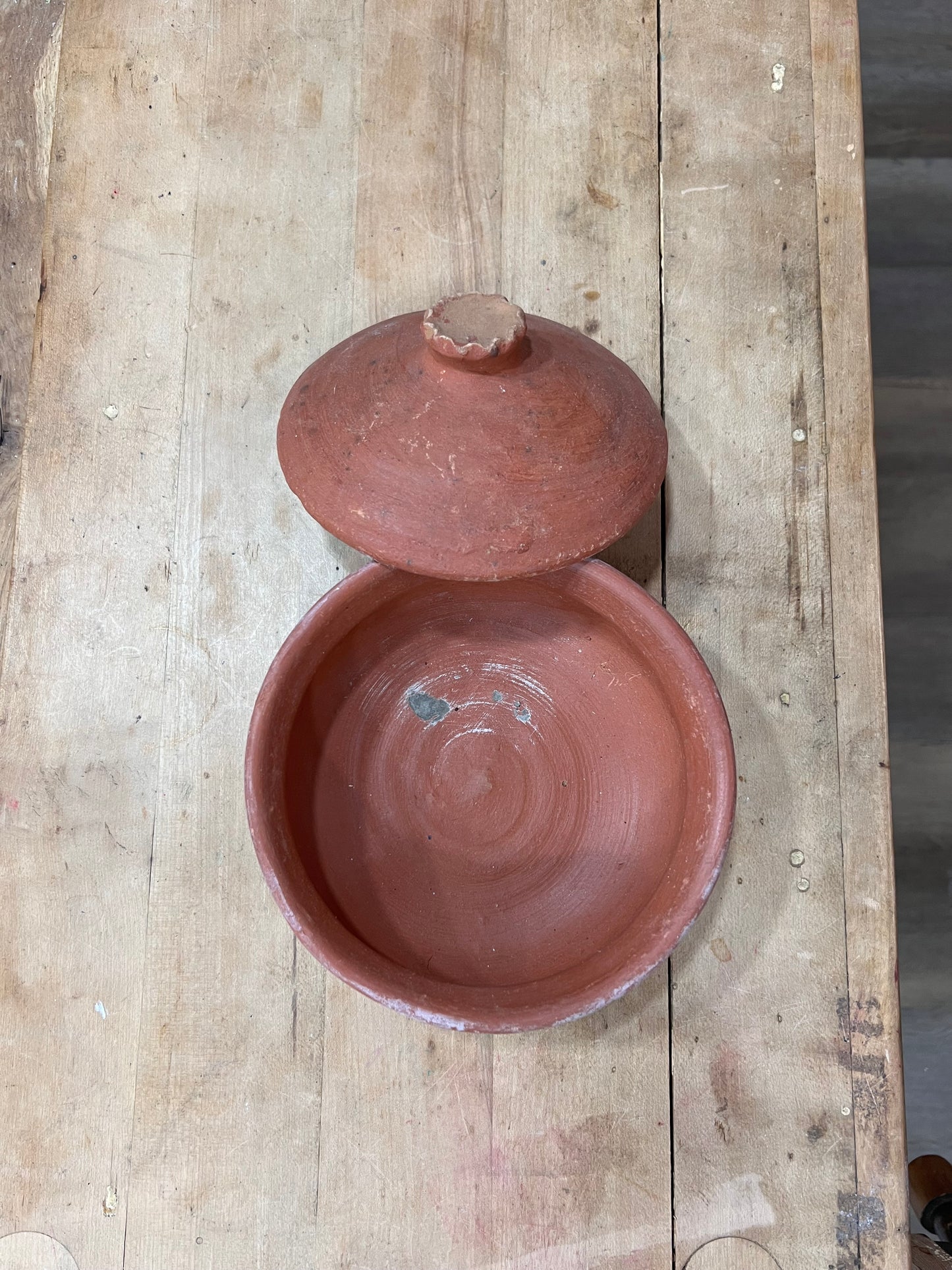 A handmade terracotta clay lidded bowl with a matching lid resting on top, displayed on a rustic wooden plank surface.
