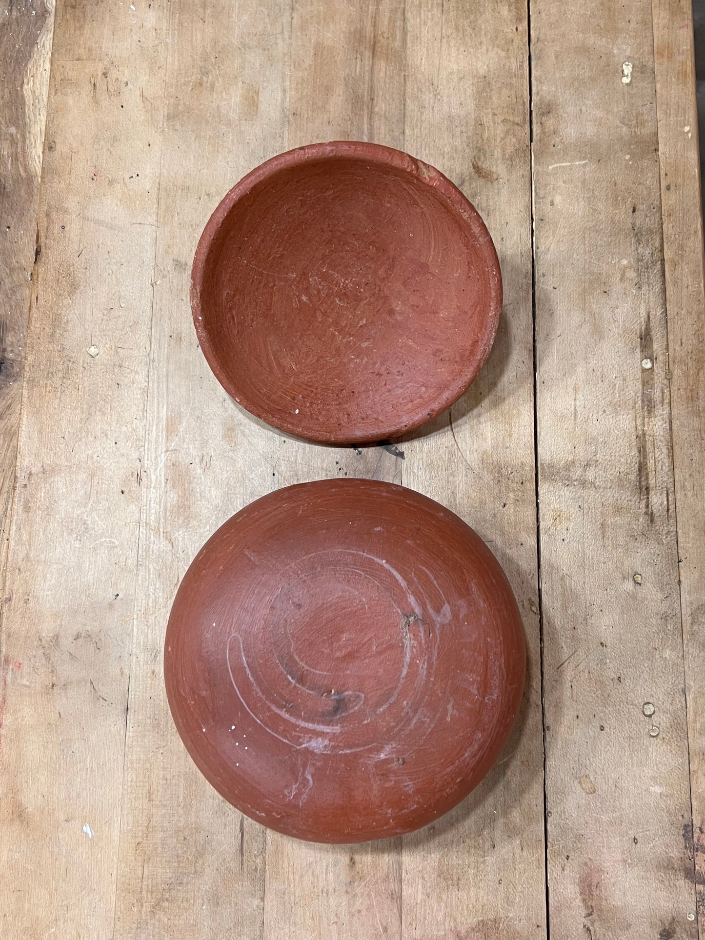 Two round terracotta clay bowls, one shown from the top and one from the bottom side, placed on a rustic wooden plank surface.
