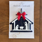 A handmade Christmas nativity scene ornament in a dark navy blue and white silhouette design, featuring a bright red bow on top, presented on its white backing card which reads 'Seed Valley Handmade Ornaments' and placed on a wooden surface.