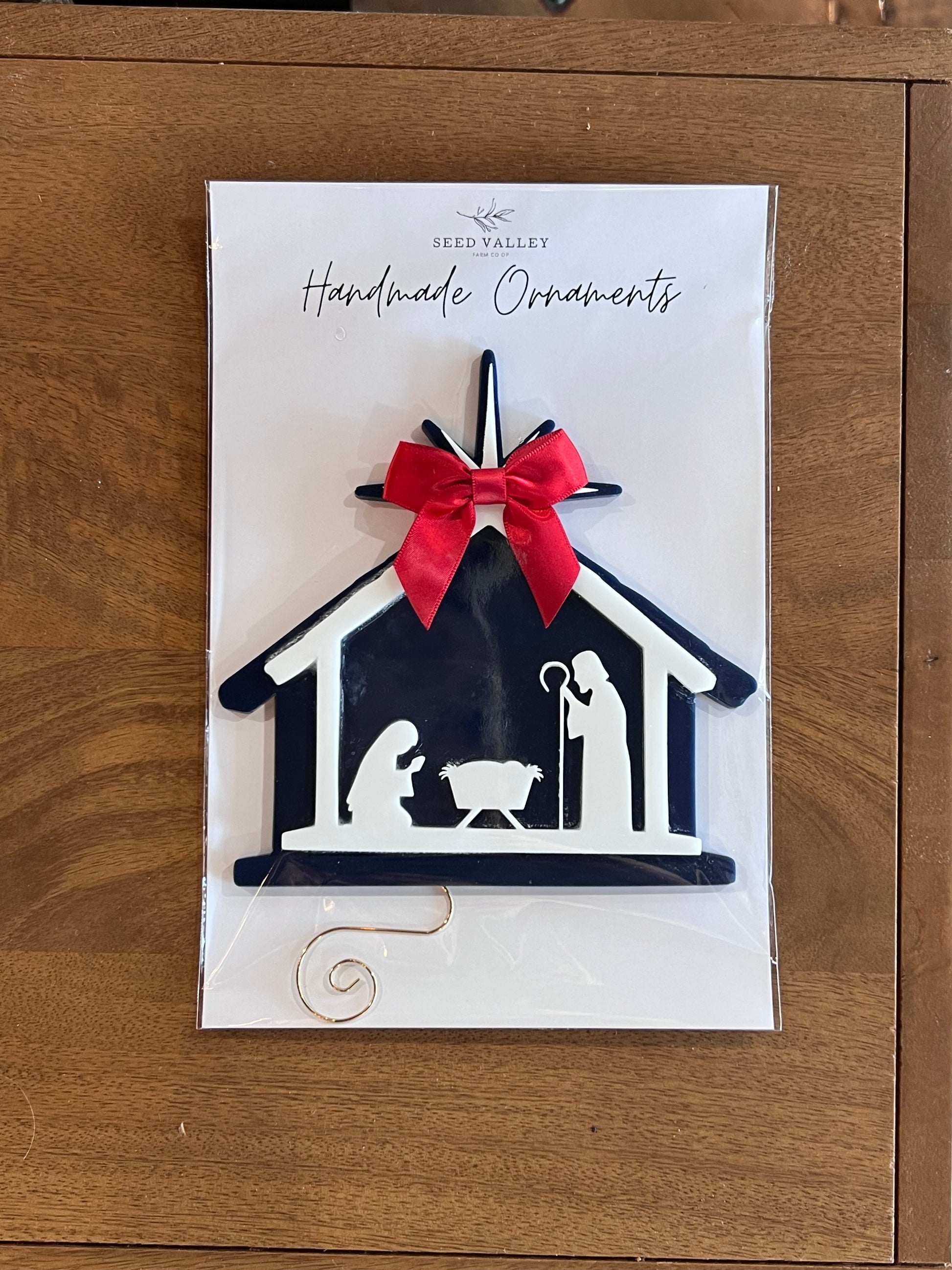 A handmade Christmas nativity scene ornament in a dark navy blue and white silhouette design, featuring a bright red bow on top, presented on its white backing card which reads 'Seed Valley Handmade Ornaments' and placed on a wooden surface.