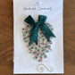 A Seed Valley handmade ornament displayed on a white backing card, featuring a scalloped green and brown patterned ornament with a dark green satin bow, placed on a wooden surface.