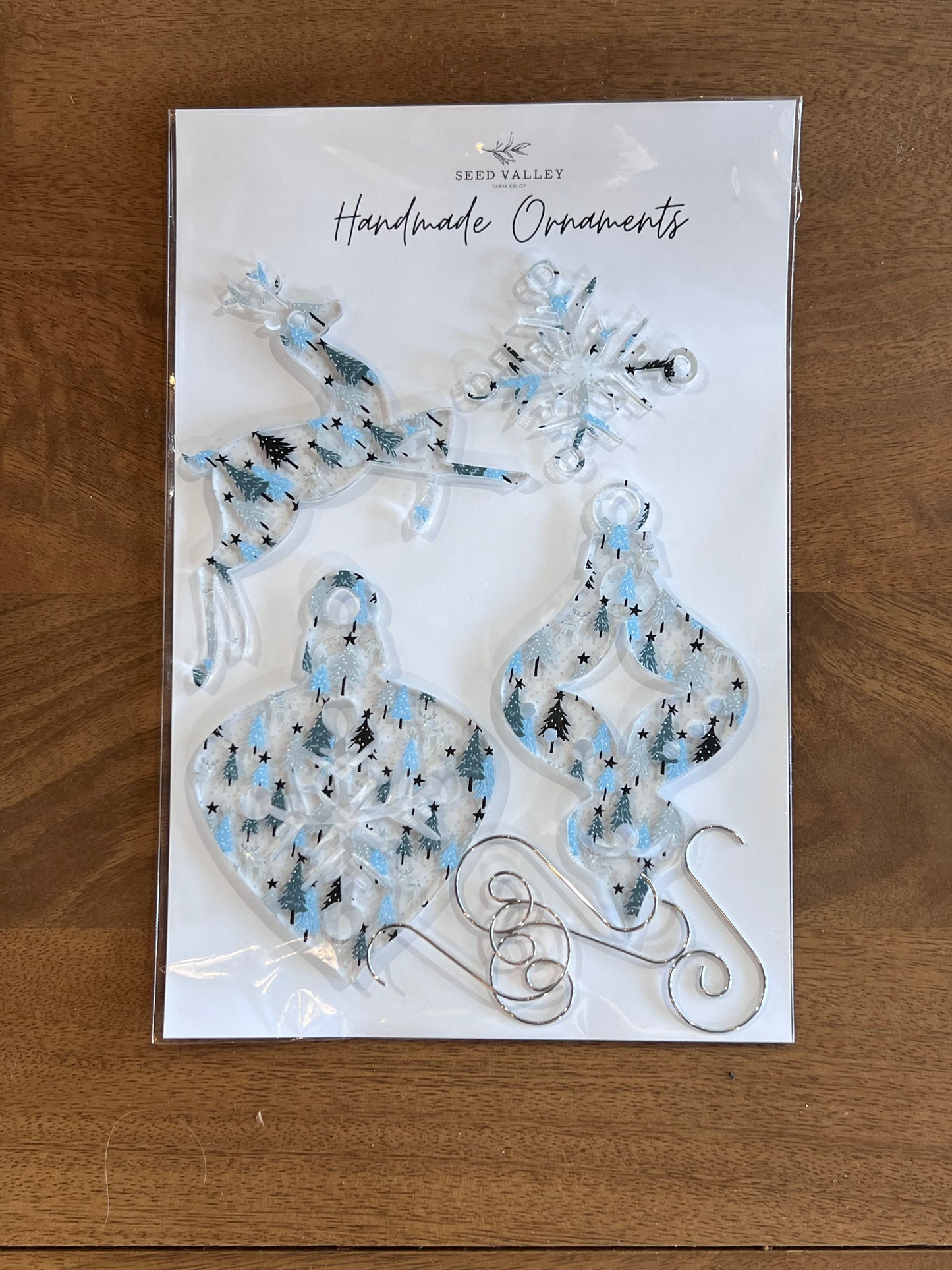 A package of Seed Valley Handmade Ornaments featuring four clear acrylic holiday ornaments with blue and black glitter patterns, displayed on a wooden table.
