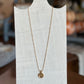 A delicate gold chain necklace with a small circular pendant displayed on a white fabric mannequin bust. The background is a softly blurred indoor setting with warm, neutral tones.
