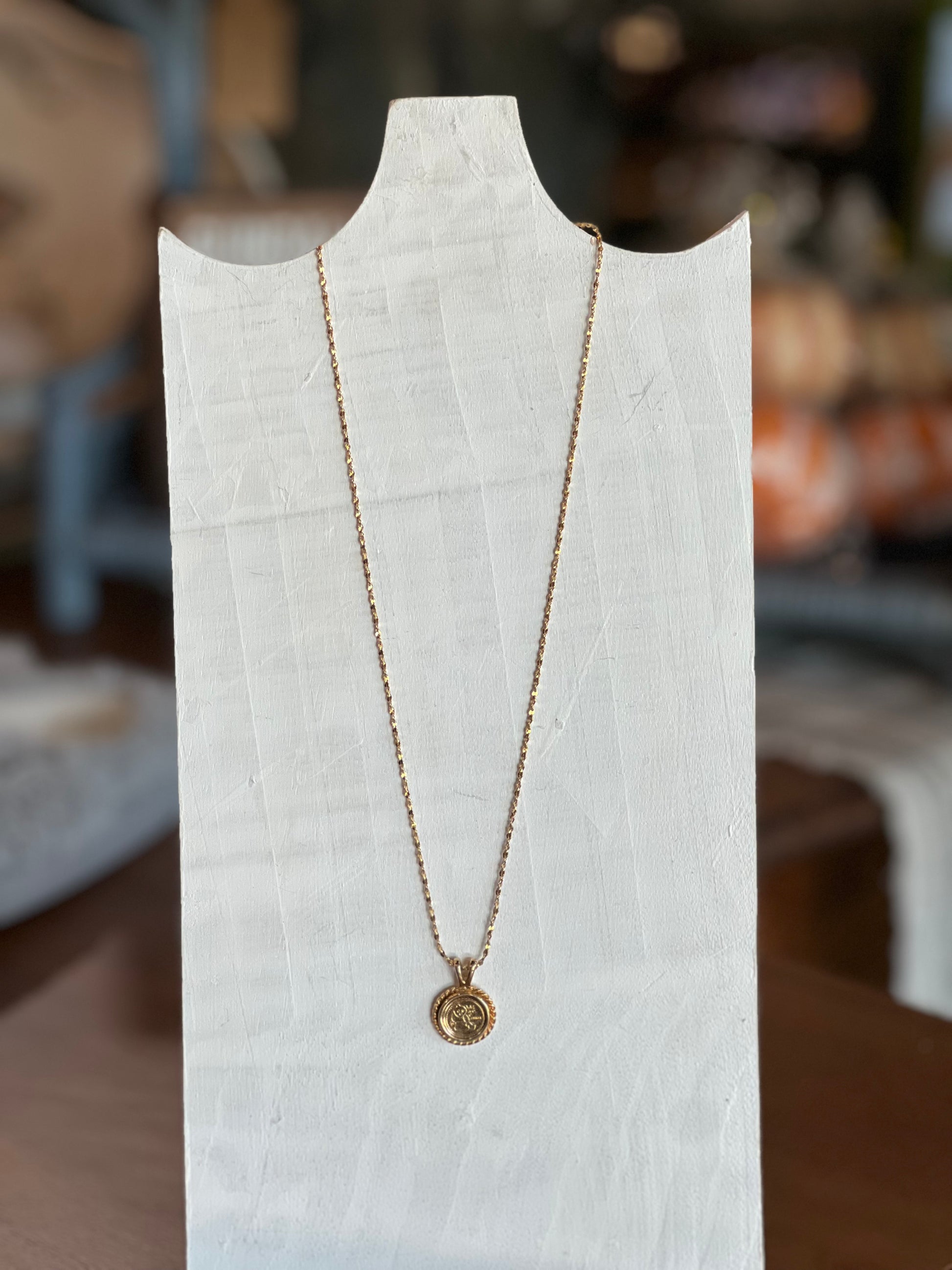 A delicate gold chain necklace with a small circular pendant displayed on a white fabric mannequin bust. The background is a softly blurred indoor setting with warm, neutral tones.