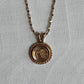 A gold necklace with a round medallion pendant featuring a floral design, displayed against a textured light gray background.