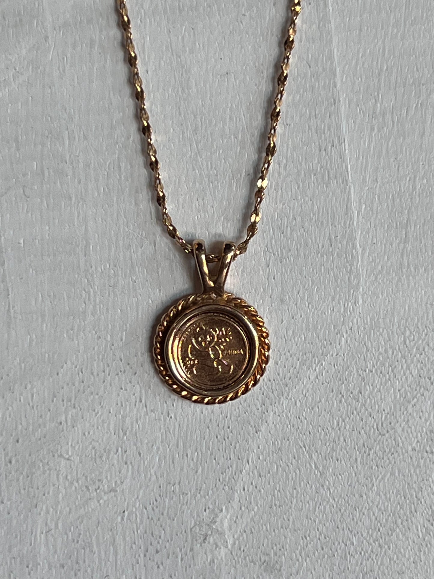 A gold necklace with a round medallion pendant featuring a floral design, displayed against a textured light gray background.