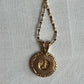 A gold pendant necklace featuring a round, textured outer border and a smaller central medallion, suspended from a twisted gold chain against a light gray background.