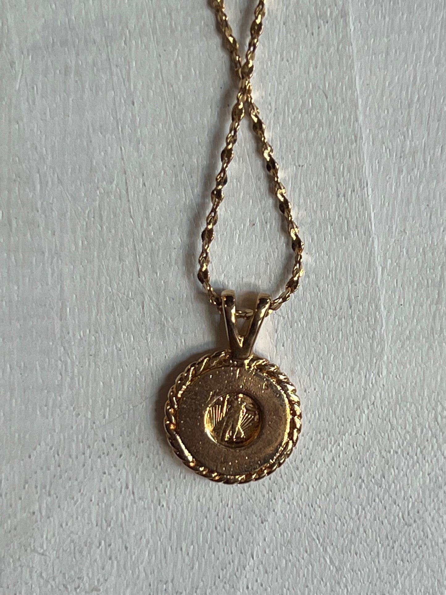 A gold pendant necklace featuring a round, textured outer border and a smaller central medallion, suspended from a twisted gold chain against a light gray background.