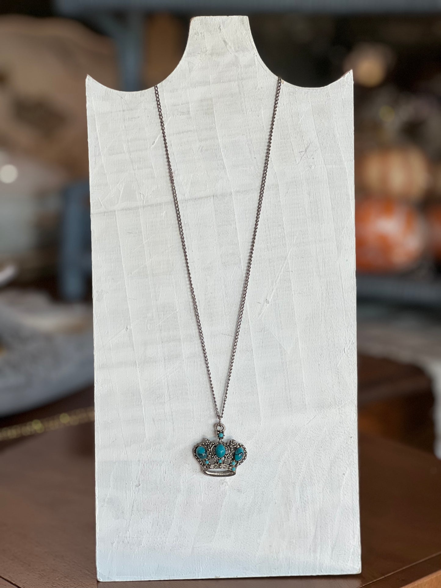 A silver crown pendant necklace with turquoise-colored stones, displayed on a white wooden stand.