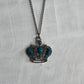 A crown-shaped pendant necklace with turquoise blue stones set in dark silver-toned metal, hanging on a matching chain against a textured white fabric background.