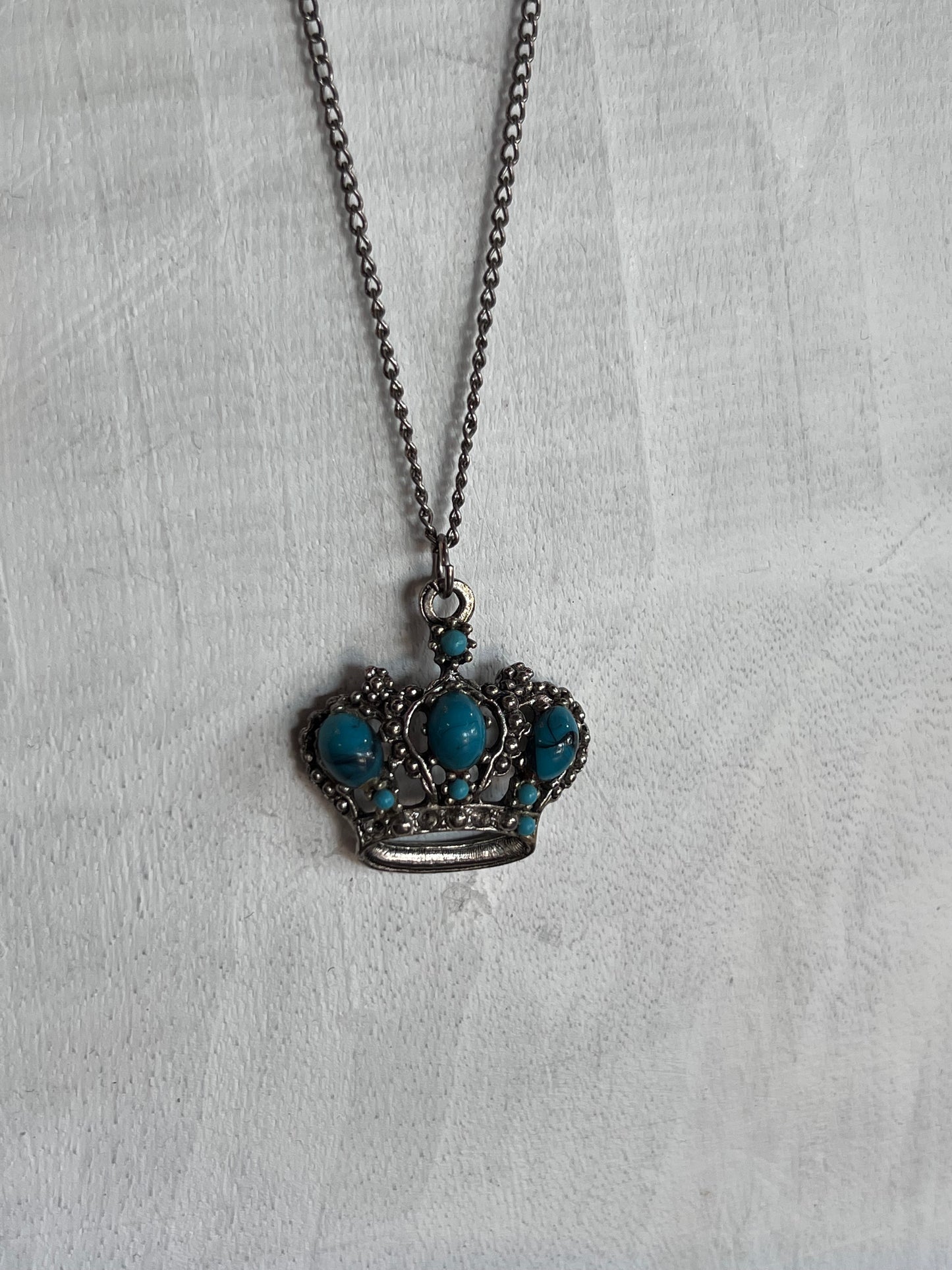 A crown-shaped pendant necklace with turquoise blue stones set in dark silver-toned metal, hanging on a matching chain against a textured white fabric background.
