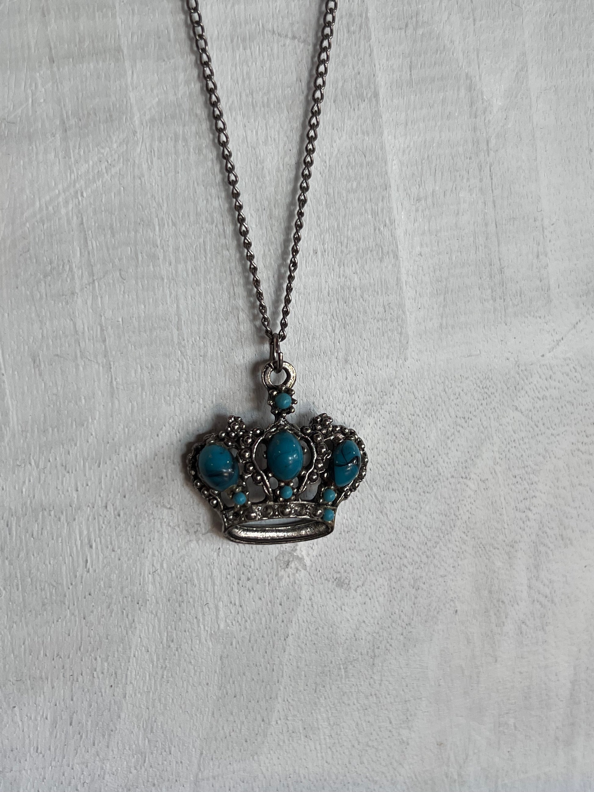 A crown-shaped pendant necklace with turquoise blue stones set in dark silver-toned metal, hanging on a matching chain against a textured white fabric background.