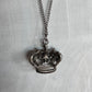 A crown-shaped pendant necklace with an oxidized silver finish, hanging from a matching chain against a textured white fabric background.