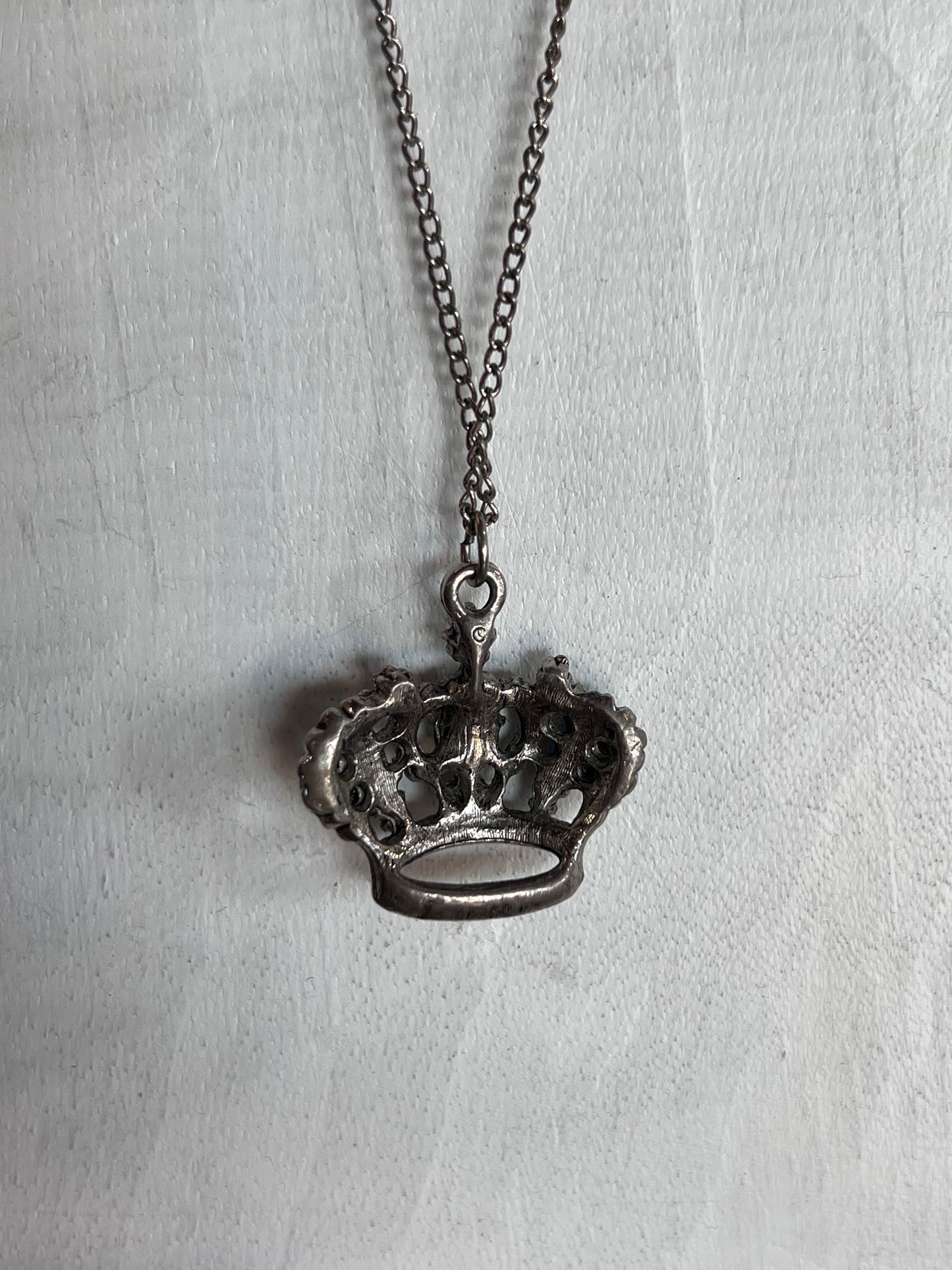 A crown-shaped pendant necklace with an oxidized silver finish, hanging from a matching chain against a textured white fabric background.
