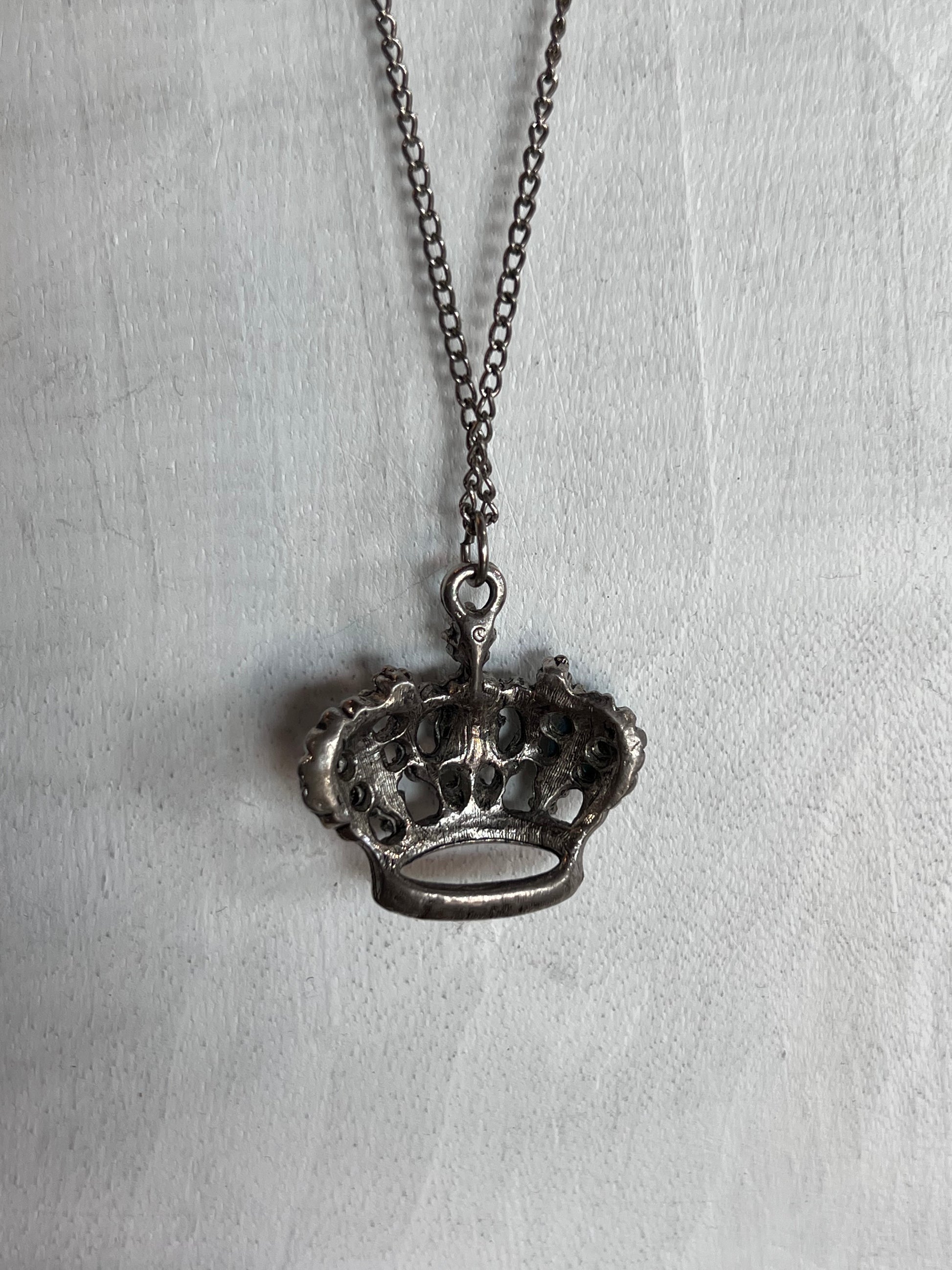 A crown-shaped pendant necklace with an oxidized silver finish, hanging from a matching chain against a textured white fabric background.