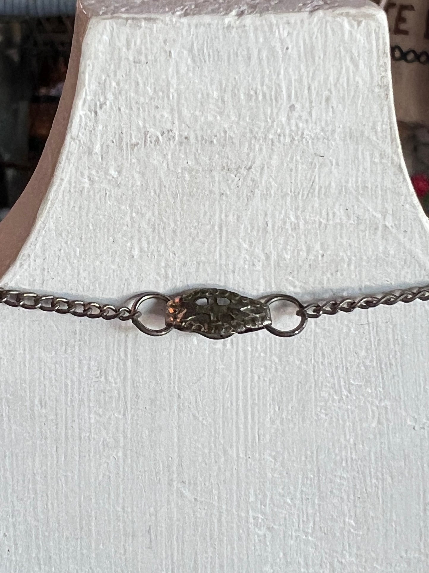 A silver chain bracelet with a textured, organic-shaped centerpiece and two small rings, displayed on a white textured bust.